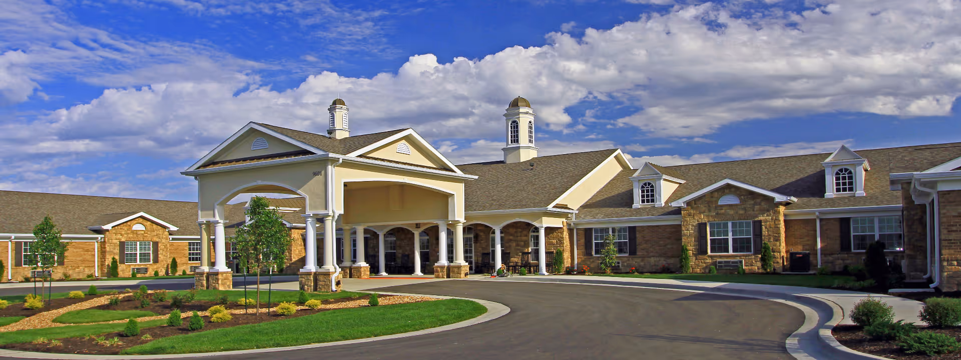 Exterior view of Benton House of Tiffany Springs, showing a large building with a covered entrance supported by white columns, stone and brick facade, landscaped garden beds, and a curved driveway under a partly cloudy blue sky.