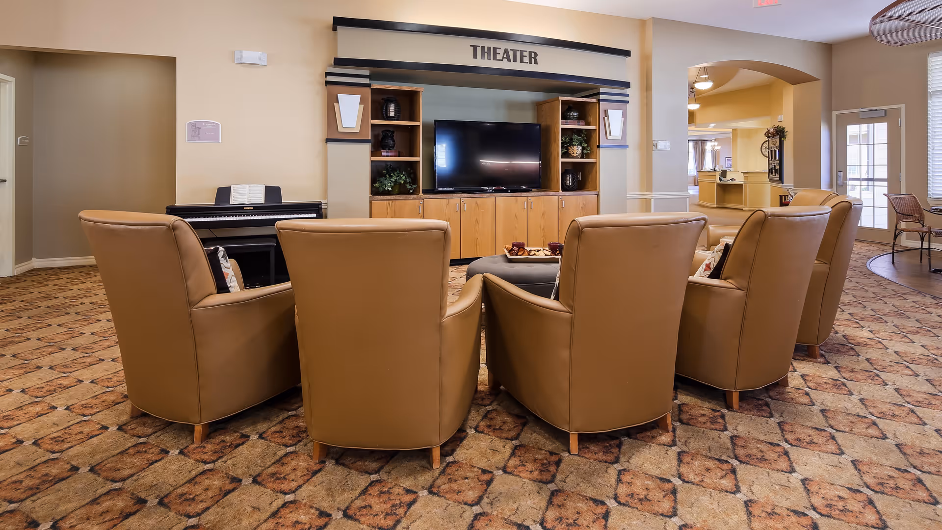 A cozy theater room in a senior living facility with six tan leather armchairs arranged in a semicircle facing a large flat-screen TV mounted on a wooden entertainment center labeled 'THEATER'. There is a piano with sheet music to the left, and the room has patterned carpet and warm beige walls.