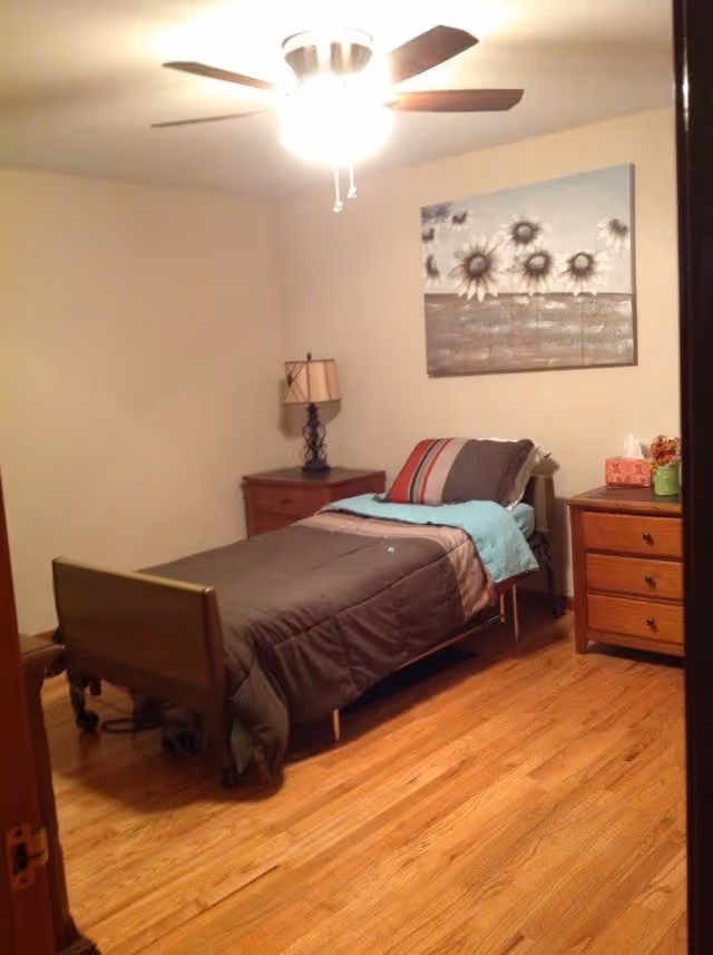 A small bedroom with a single bed covered in a brown comforter and a blue blanket. There are two wooden nightstands on either side of the bed, one with a lamp and the other with a tissue box and a small plant. A ceiling fan with a light is on, and a painting of flowers hangs on the wall above the bed. The floor is wooden.