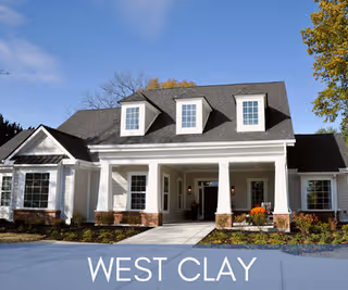 Front entrance of a single-story white cottage-style senior living building with a covered porch and three dormer windows under a blue sky.