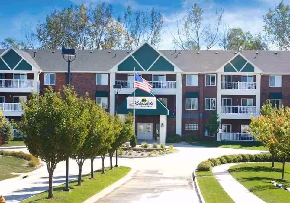 Exterior view of The Arbordale senior living building with trees, walkway, and flag