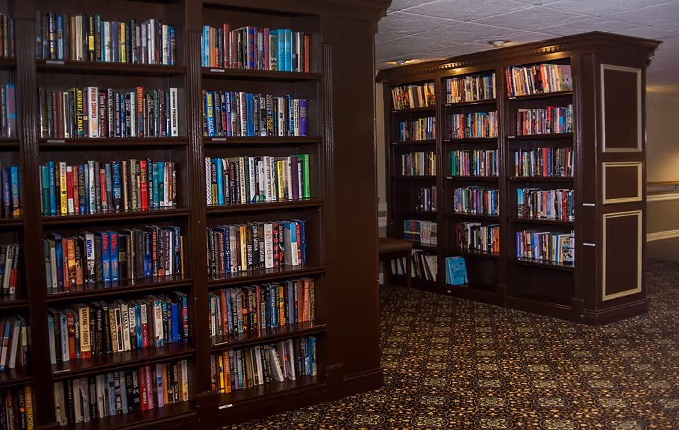 Interior view of a library area in an assisted living facility with dark wooden bookshelves filled with books and a patterned carpeted floor.