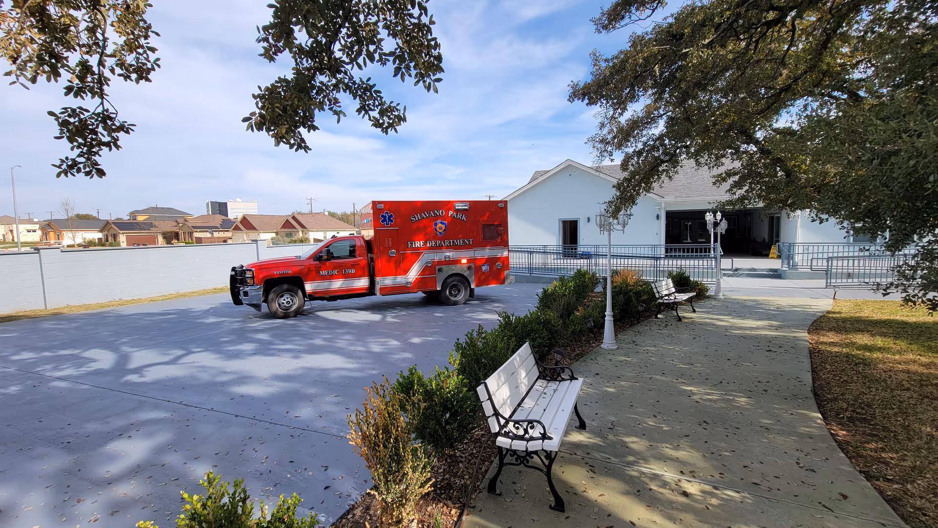 Outdoor view of a facility with a paved area and benches along a walkway. A red Shavano Park Fire Department ambulance is parked on the paved area near a white building with a ramp entrance. Trees and bushes are visible around the walkway.