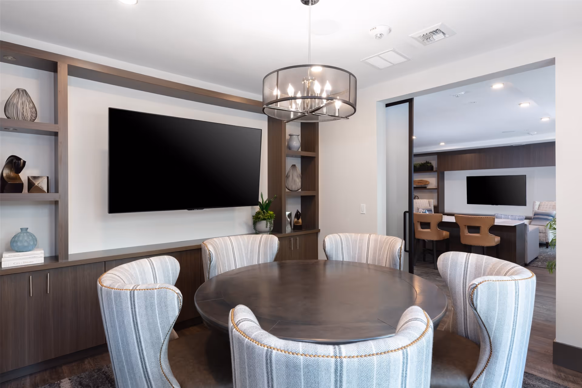 A modern interior room featuring a round dark wood table surrounded by six upholstered chairs with striped fabric. On the wall behind the table is a large flat-screen TV mounted above built-in wooden shelves and cabinets decorated with vases and books. A contemporary chandelier hangs above the table. In the background, another room is visible with additional seating and another mounted TV.