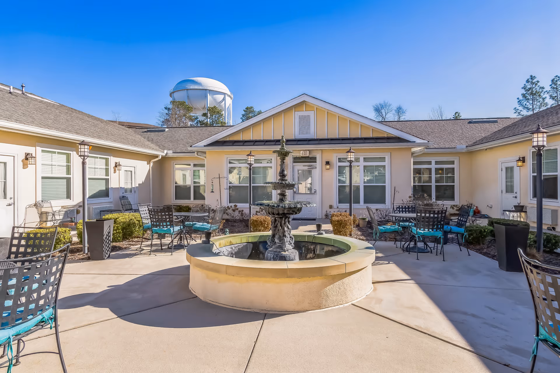 Outdoor courtyard area of a senior living facility with a central water fountain surrounded by metal chairs with teal cushions and tables. The courtyard is enclosed by a single-story building with beige walls and multiple windows and doors. A water tower and trees are visible in the background under a clear blue sky.