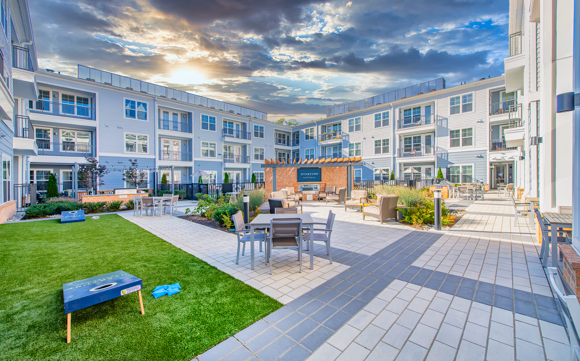 Outdoor courtyard area of Overture Centennial senior living facility featuring a green lawn with cornhole boards, multiple seating areas with tables and chairs, a pergola with a fireplace, and a multi-story building surrounding the courtyard under a partly cloudy sky at sunset.