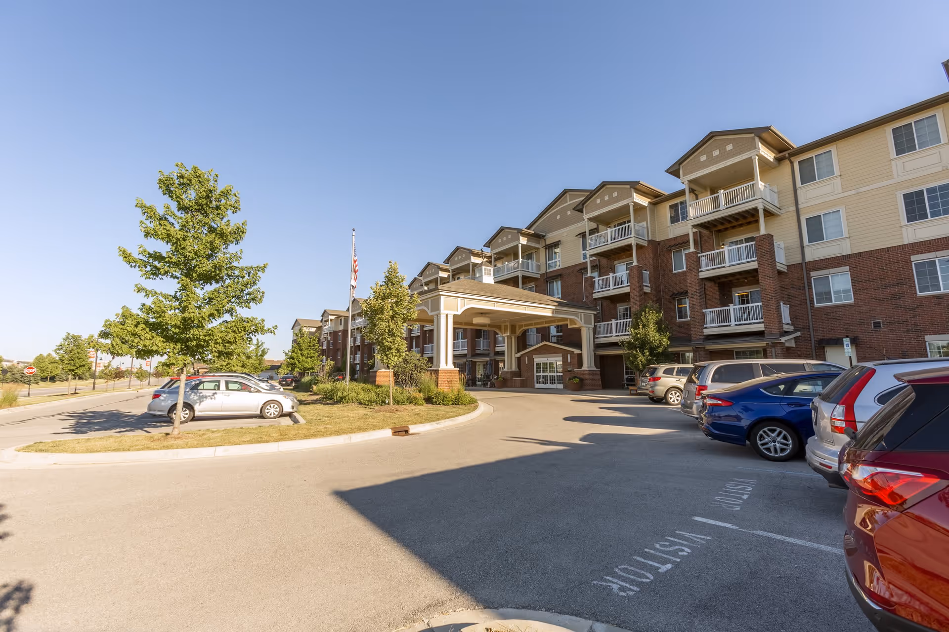 Exterior view of Independence Village of Zionsville West, showing a multi-story senior living building with balconies, a covered entrance, parked cars, trees, and a clear blue sky.