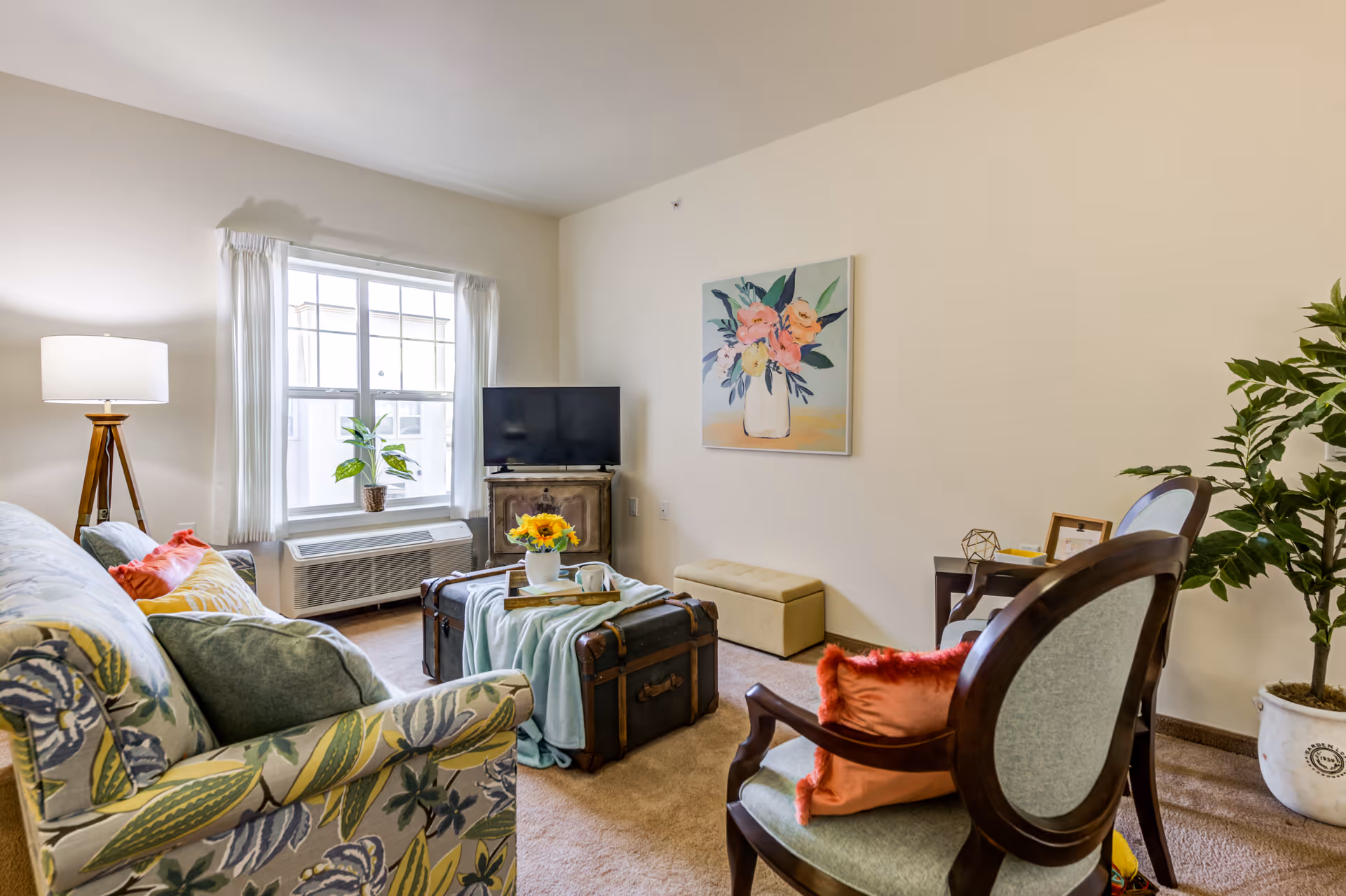 A cozy living room with a patterned sofa and armchairs around a trunk coffee table, a TV by the window, and floral wall art.