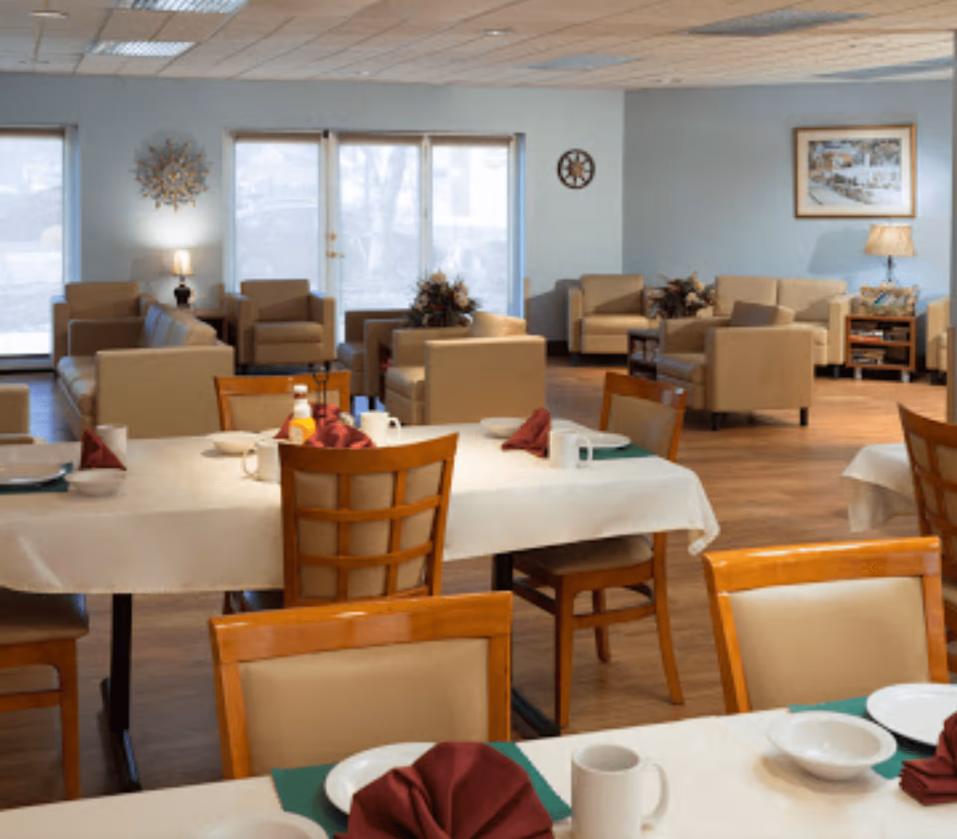 A spacious dining and lounge area in a senior living facility with tables set with white tablecloths, plates, bowls, mugs, and folded burgundy napkins. The background features beige armchairs and sofas arranged around coffee tables, with lamps, wall art, and large windows letting in natural light.
