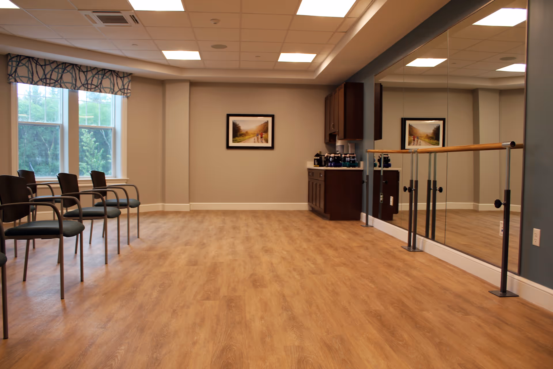 A spacious room with wooden flooring and a row of chairs along the left wall under two large windows with patterned valances. The right wall features a large mirror with a ballet barre in front of it. At the far end of the room, there is a countertop with cabinets above and below, holding several water bottles. A framed landscape picture hangs on the back wall.