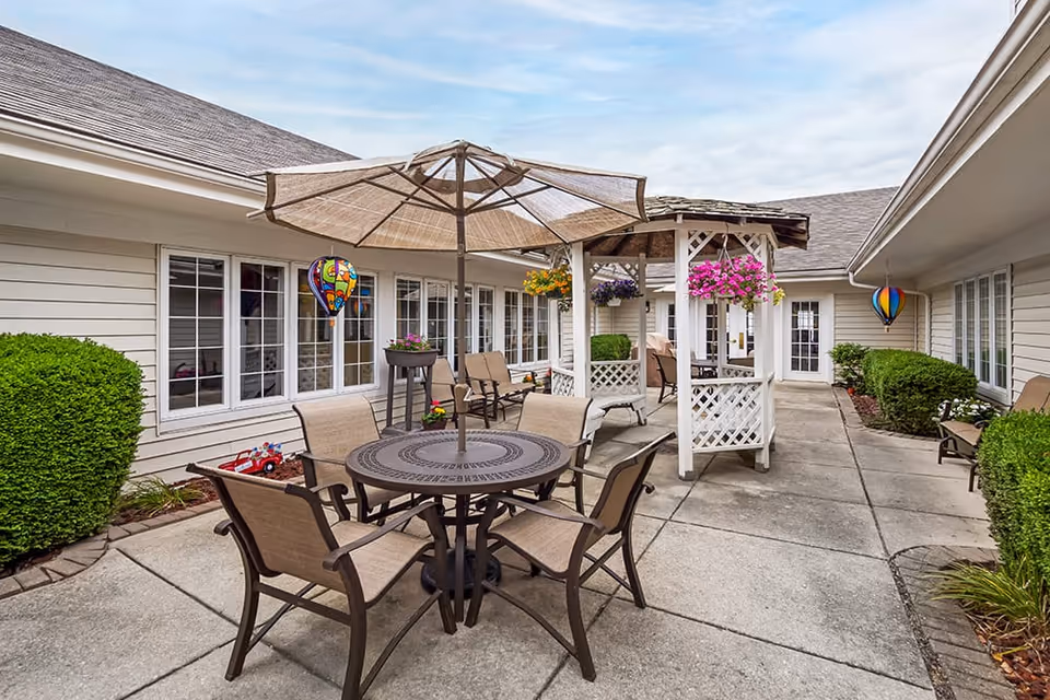 Outdoor patio area at Brookdale Farmington Hills North featuring a round metal table with four beige chairs and a large beige umbrella. There is a white wooden gazebo adorned with hanging flower baskets and additional seating. The patio is surrounded by beige siding buildings with multiple windows and green bushes along the edges. Colorful hot air balloon decorations hang near the windows.