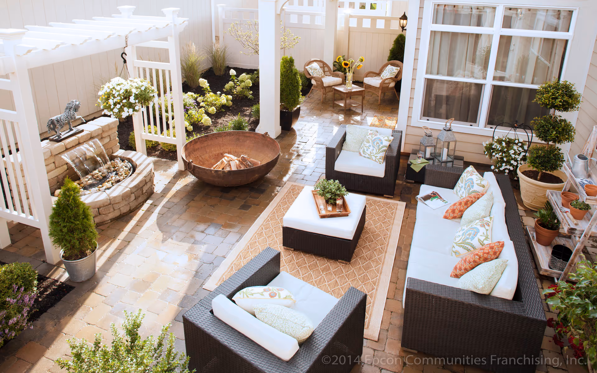 A cozy outdoor patio area with wicker furniture including a sofa, two armchairs, and an ottoman with cushions and decorative pillows. There is a small table with a vase of sunflowers and two chairs in the background. The patio features a water fountain with a horse statue, a fire pit, potted plants, and a pergola with hanging flower baskets. The area is paved with stone tiles and surrounded by garden beds with flowers and shrubs.