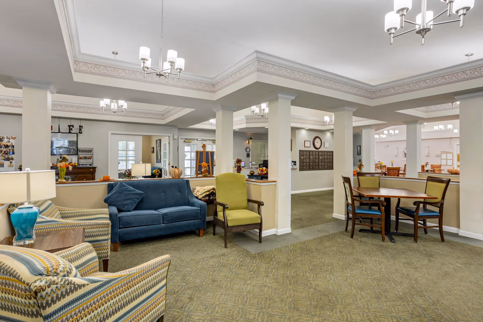 A spacious and well-lit common area in a senior living facility featuring comfortable seating including a blue sofa, green armchair, and patterned armchairs. There are multiple tables and chairs for dining or socializing, decorative lamps, and ceiling lights. The room has white walls with decorative molding and carpeted floors. In the background, there is a wall with mailboxes and a clock.