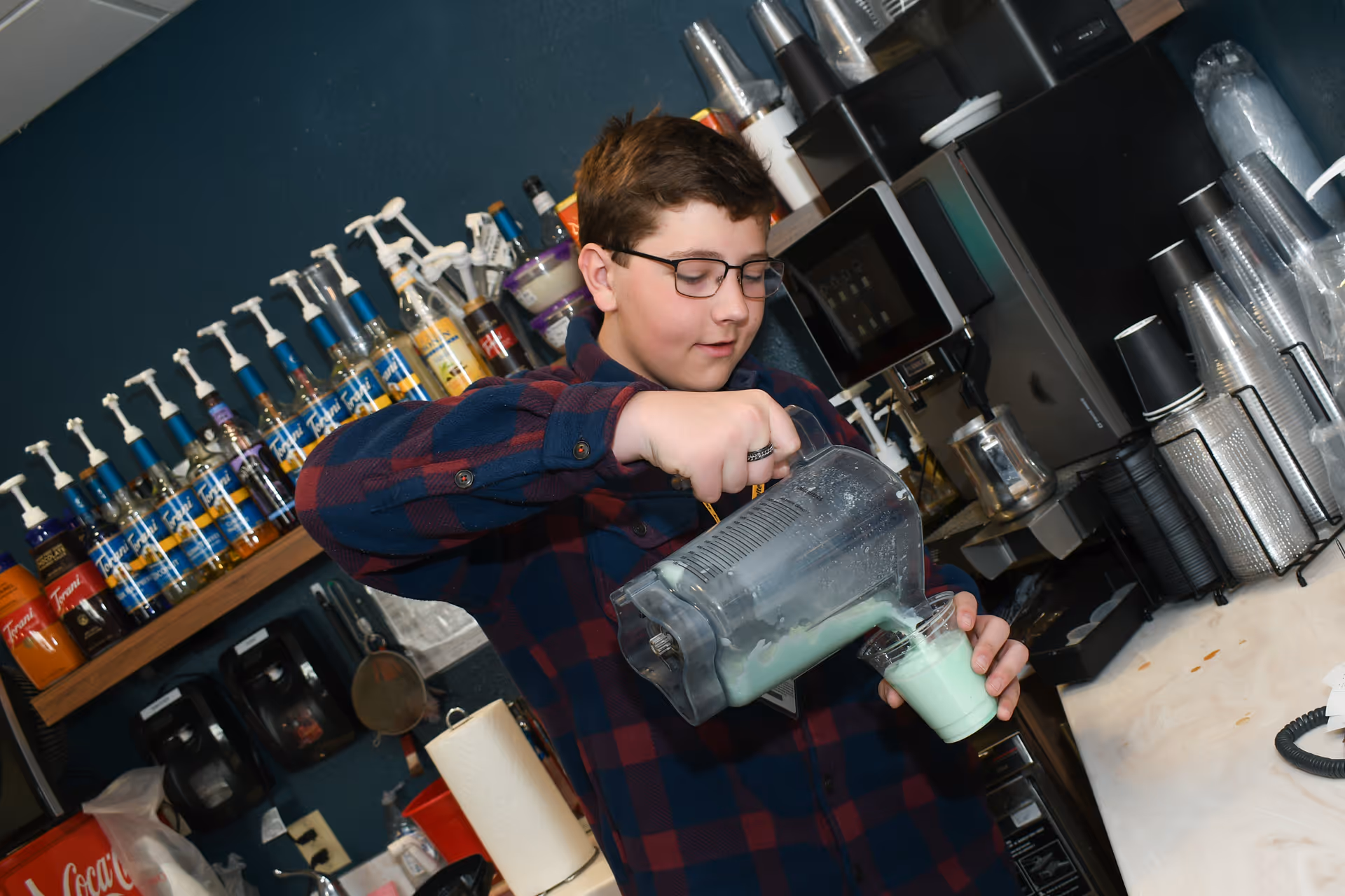A young person wearing glasses and a plaid shirt is pouring a green smoothie or drink from a blender pitcher into a clear plastic cup in a kitchen or beverage preparation area with various syrup bottles and a coffee machine in the background.