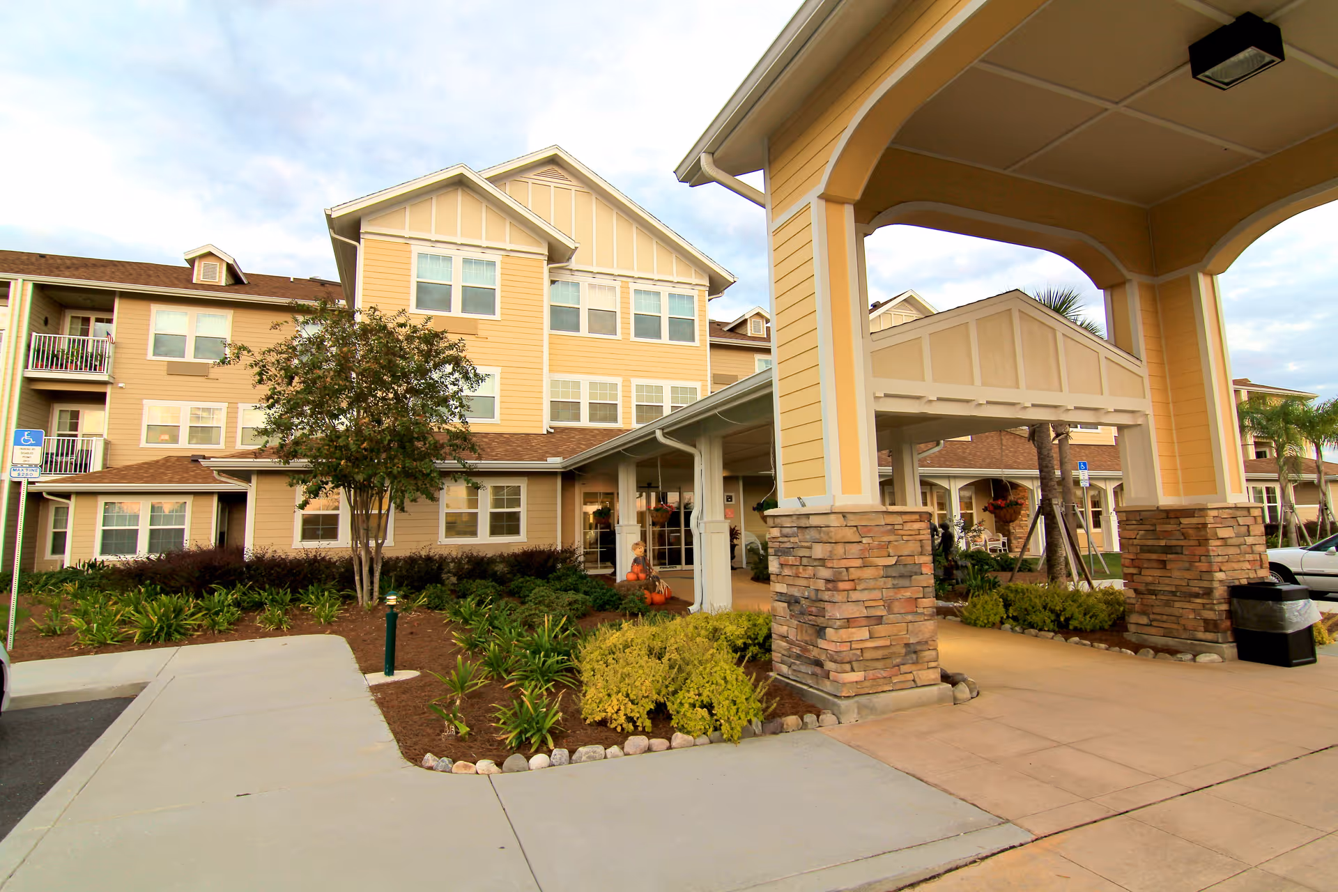 Exterior view of a multi-story senior living facility building with beige siding and stone pillars at the entrance. There are landscaped bushes and plants along the walkway leading to the entrance, with a covered drop-off area supported by stone columns. The sky is partly cloudy.