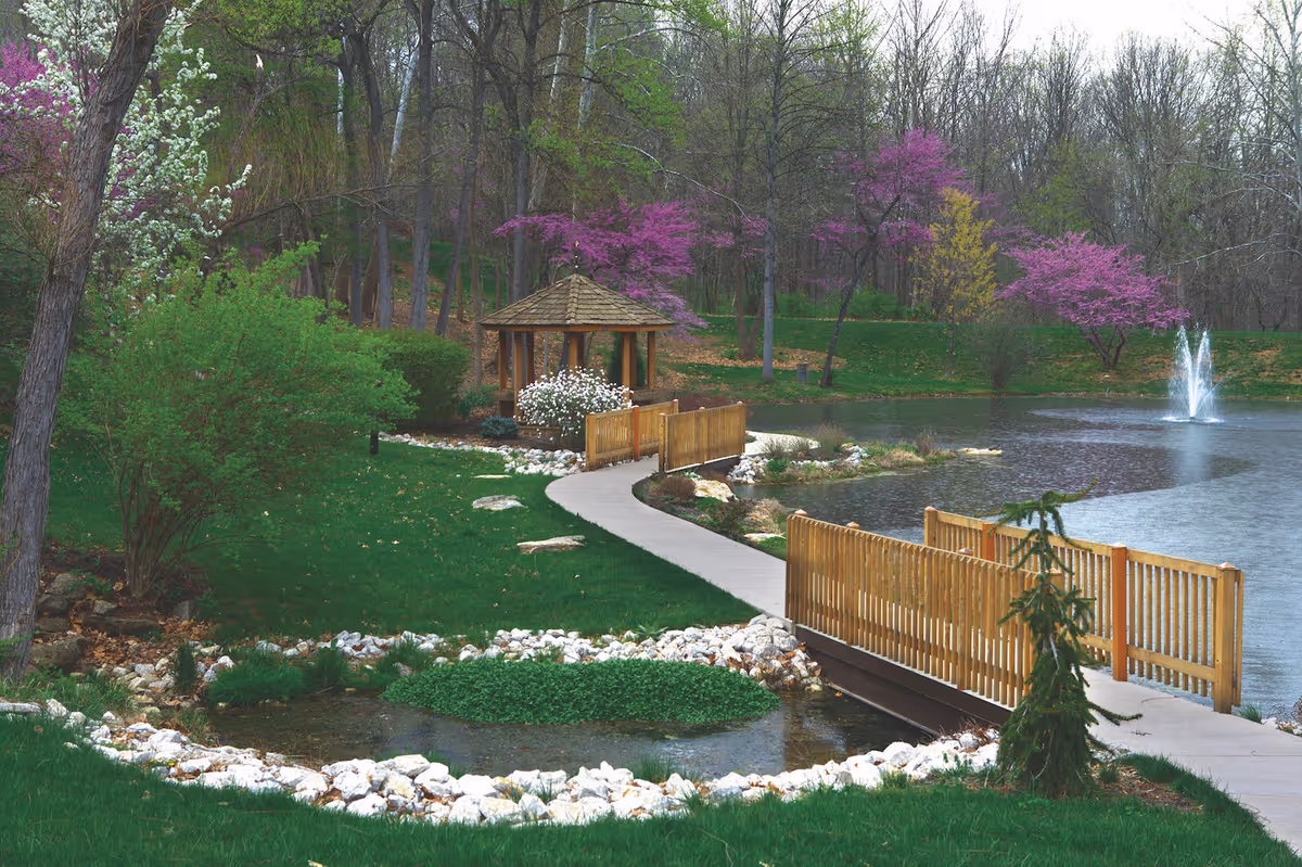 A peaceful outdoor scene featuring a wooden bridge over a small pond leading to a gazebo surrounded by green grass, blooming trees with white and purple flowers, and a water fountain in a larger pond in the background.