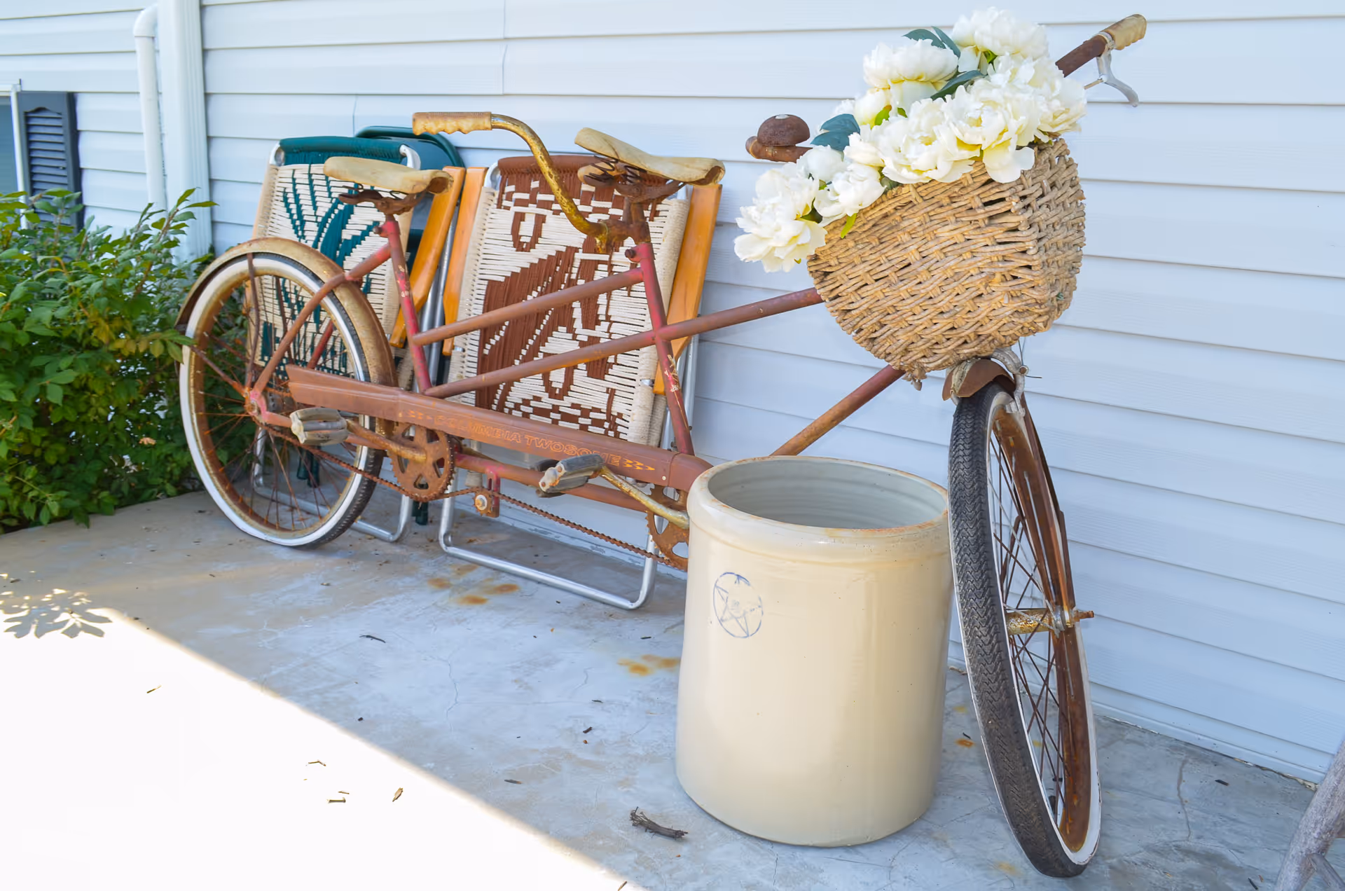A vintage tandem bicycle with a wicker basket filled with white flowers attached to the front, parked on a concrete porch next to a large ceramic crock and some colorful woven chairs, against the backdrop of white siding and green shrubbery.