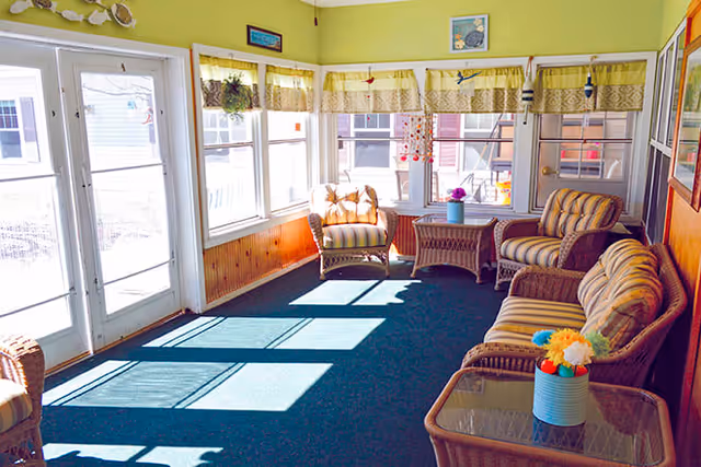 A sunlit sitting area with wicker furniture including a sofa, armchairs, and glass-top tables. The room has large windows with yellow and white curtains, green walls, and a blue carpet. There are decorative items like flowers and hanging ornaments.