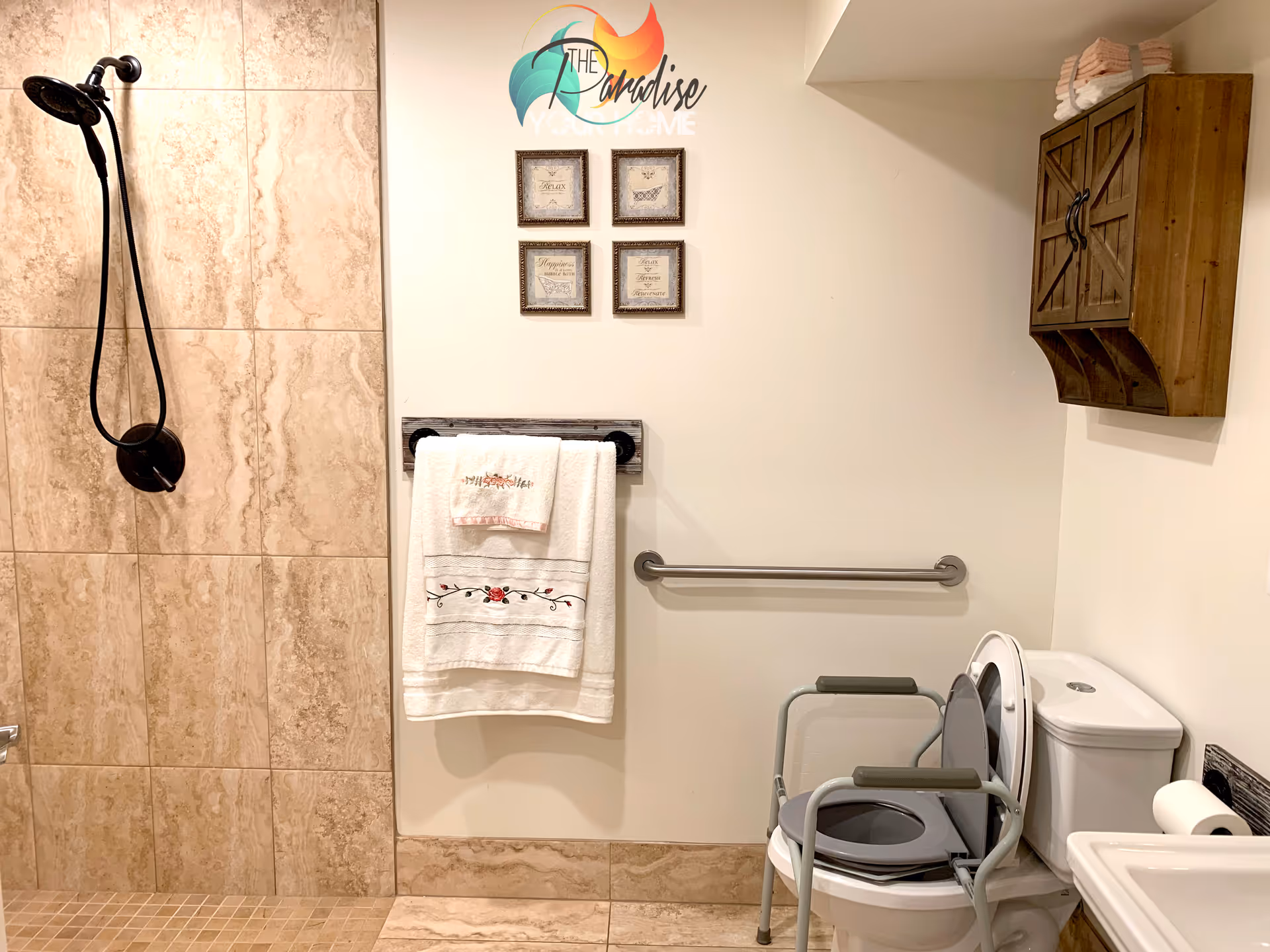A bathroom with beige tiled shower area on the left, a towel rack with two white towels hanging on the wall, a toilet with a raised seat and armrests, a wooden cabinet mounted on the wall, and a grab bar for support.