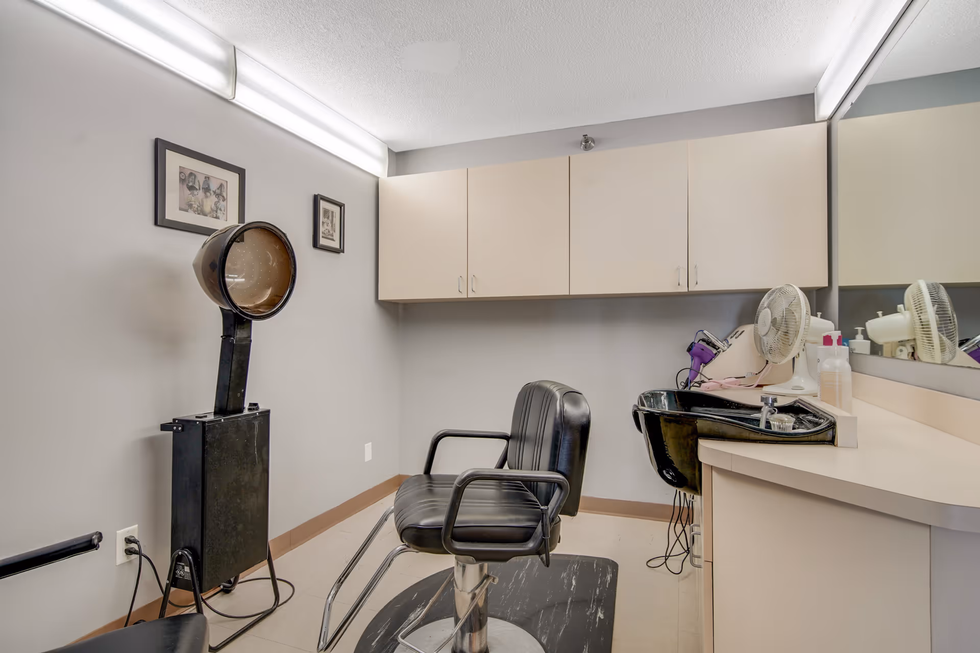 Interior of a small hair salon or beauty station with a black salon chair, a hair dryer hood, a black sink with a faucet, a countertop with a fan and hair styling tools, beige cabinets mounted on the wall, and two framed pictures hanging on a light gray wall.