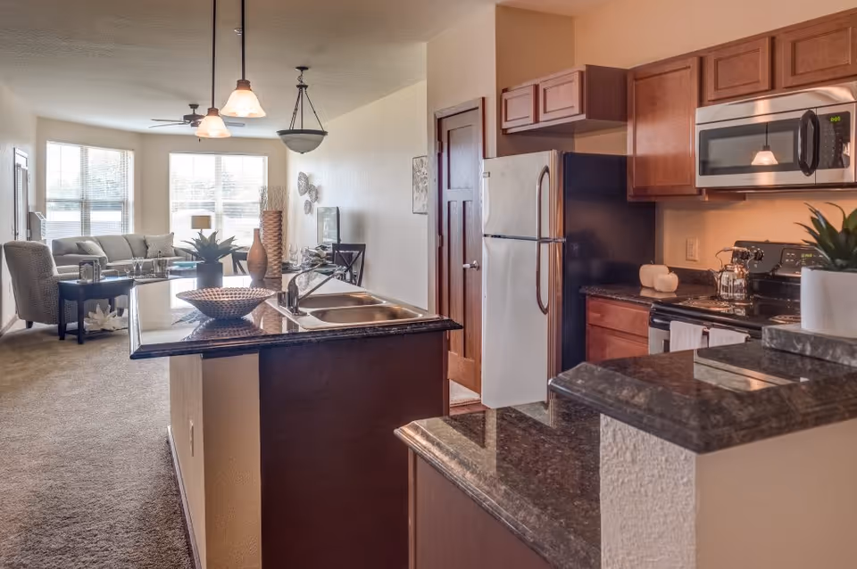 Interior view of a senior living apartment showing a kitchen with dark granite countertops, wooden cabinets, a refrigerator, microwave, stove, and a double sink. The kitchen opens into a living room area with a sofa, armchair, coffee table, and large windows letting in natural light.