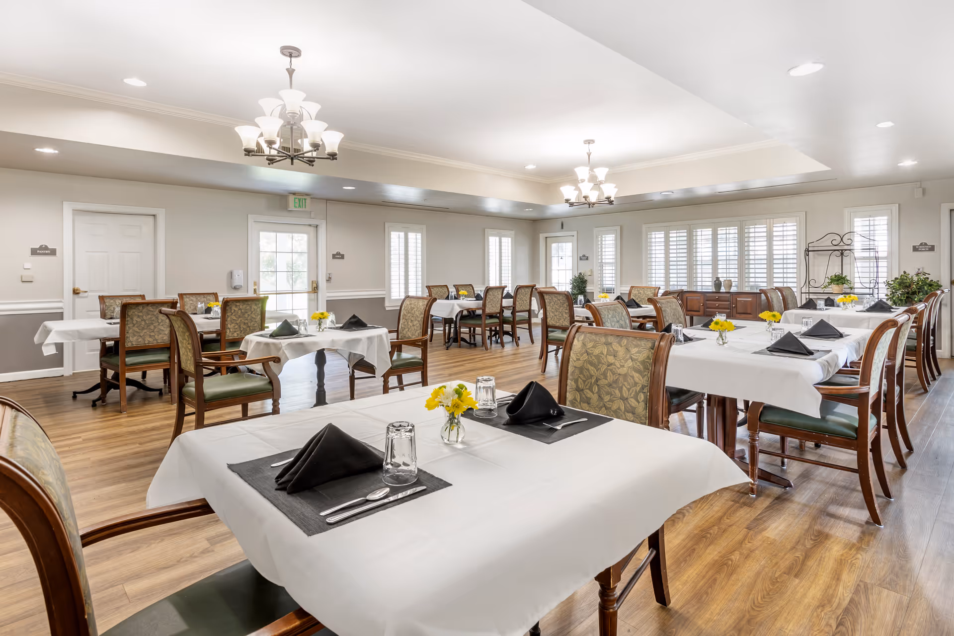 Bright dining room with multiple tables set with white tablecloths, black napkins, small flower vases, and wooden chairs.
