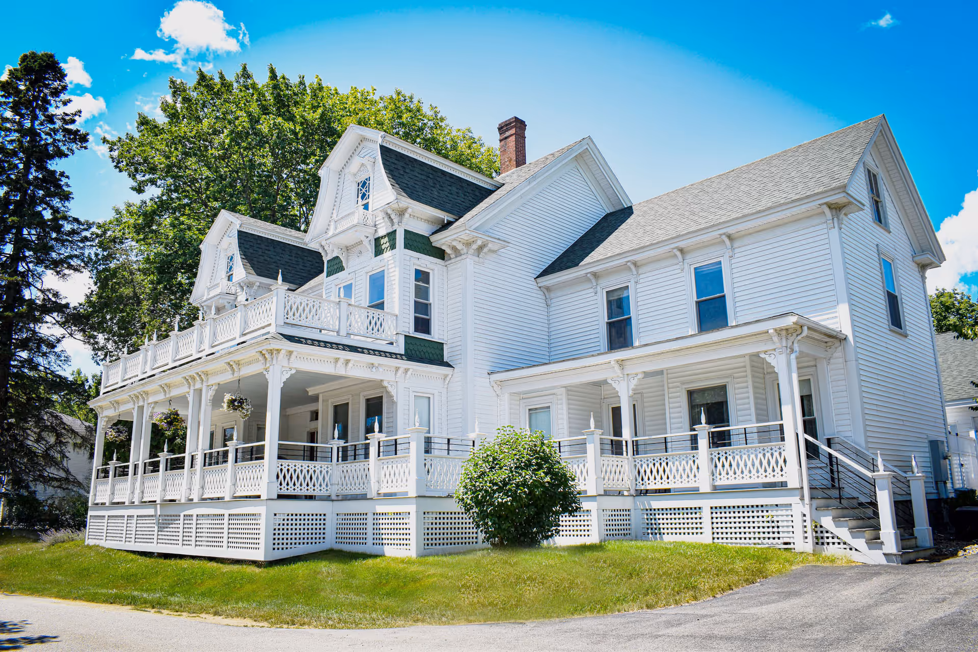 A large white Victorian-style multi-story house with a wraparound porch, decorative trim, and a small lawn under a bright blue sky.