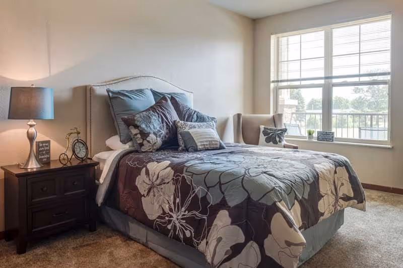 A cozy bedroom with a neatly made bed featuring floral patterned bedding and multiple pillows. Next to the bed is a dark wooden nightstand with a lamp, a small decorative bicycle, and a clock. A large window with blinds lets in natural light, and there is a cushioned armchair with a pillow near the window.