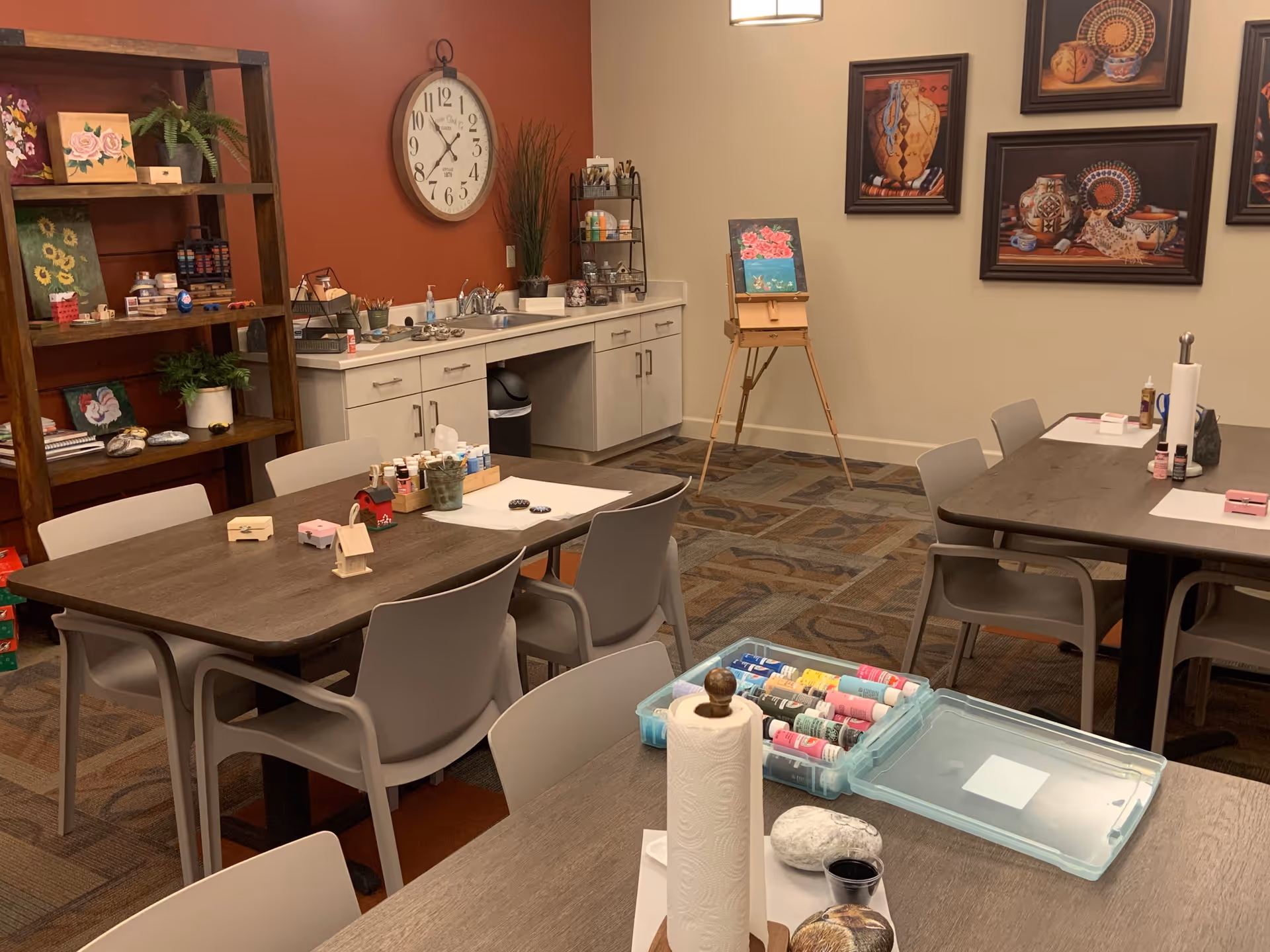 A well-lit art and craft room with several tables and chairs. The tables have various art supplies including paint bottles, brushes, paper towels, and painted rocks. There is a wooden shelf with plants and art supplies, a wall clock, a small sink area with cabinets, and an easel displaying a painting. The walls have framed artwork featuring pottery and decorative items.