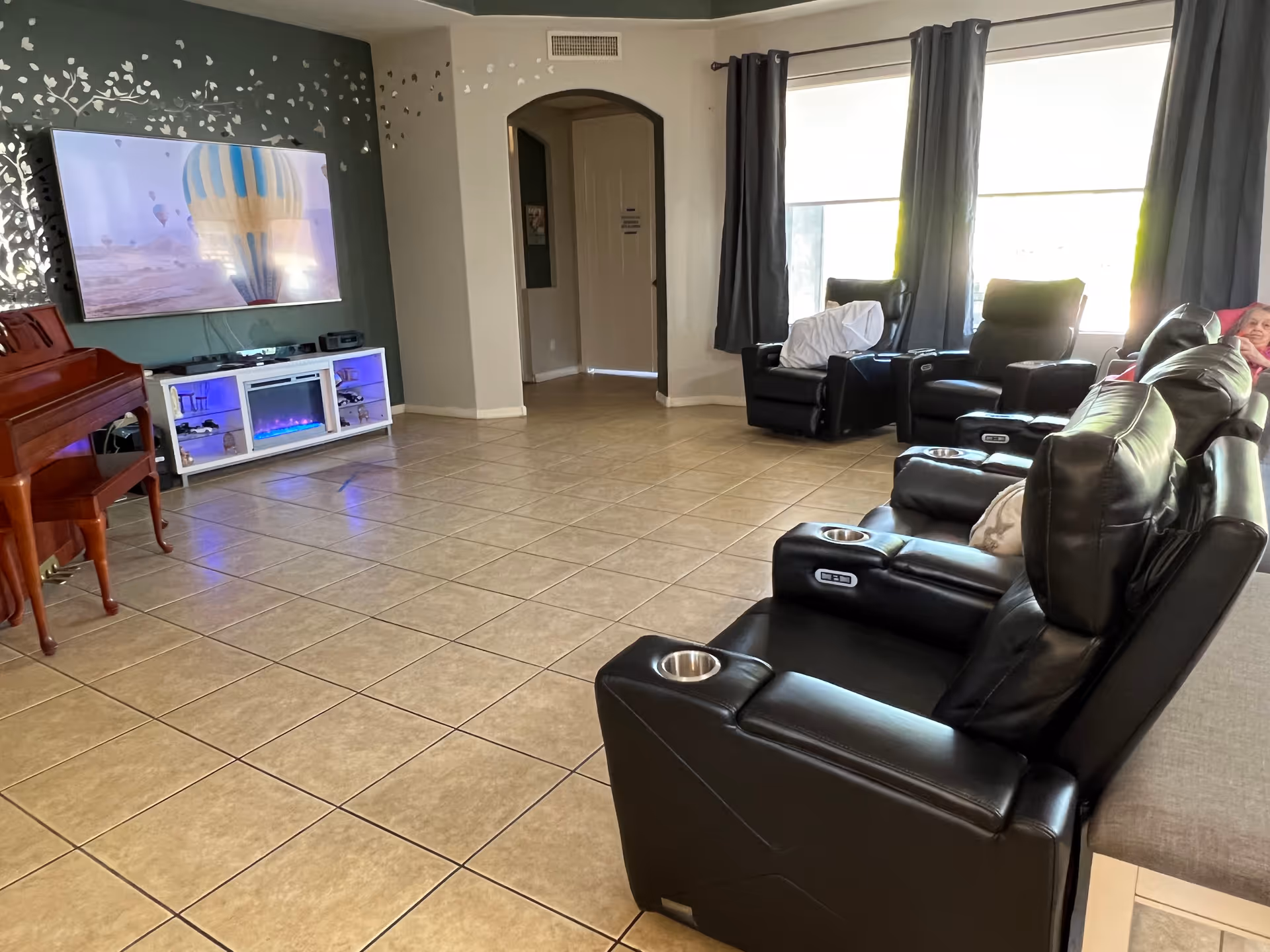A spacious living room with tiled floors, featuring a row of black leather recliners with cup holders facing a wall-mounted flat-screen TV displaying a hot air balloon scene. There is a white entertainment unit with blue lighting and a small electric fireplace below the TV. A wooden piano is positioned on the left side of the room. Large windows with dark curtains allow natural light to brighten the space.