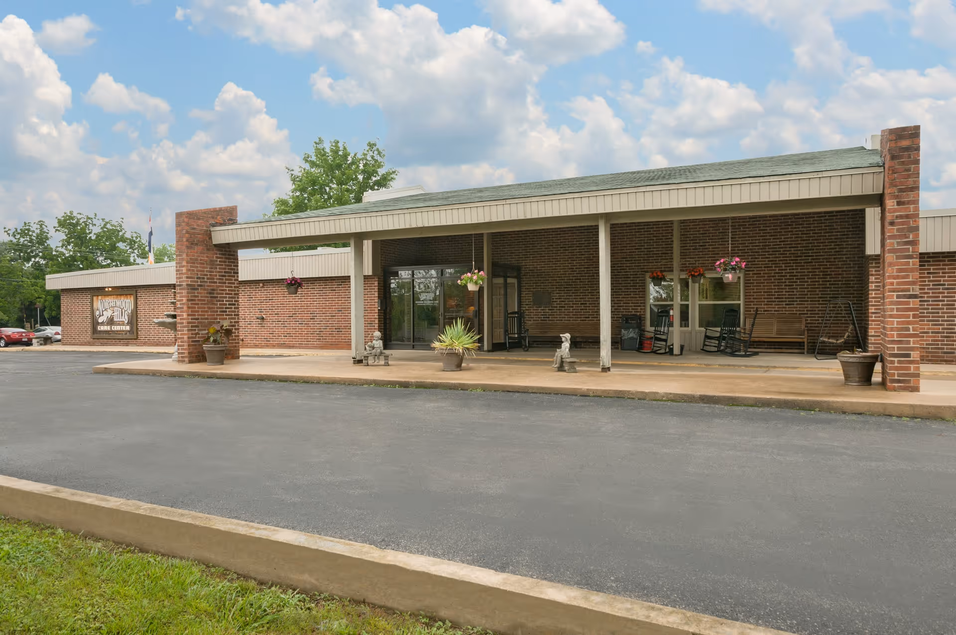 Front exterior view of Northwood Hills Care Center, a single-story brick building with a covered entrance supported by white pillars. There are hanging flower pots, potted plants, rocking chairs, and small statues on the porch area. The sky is partly cloudy.