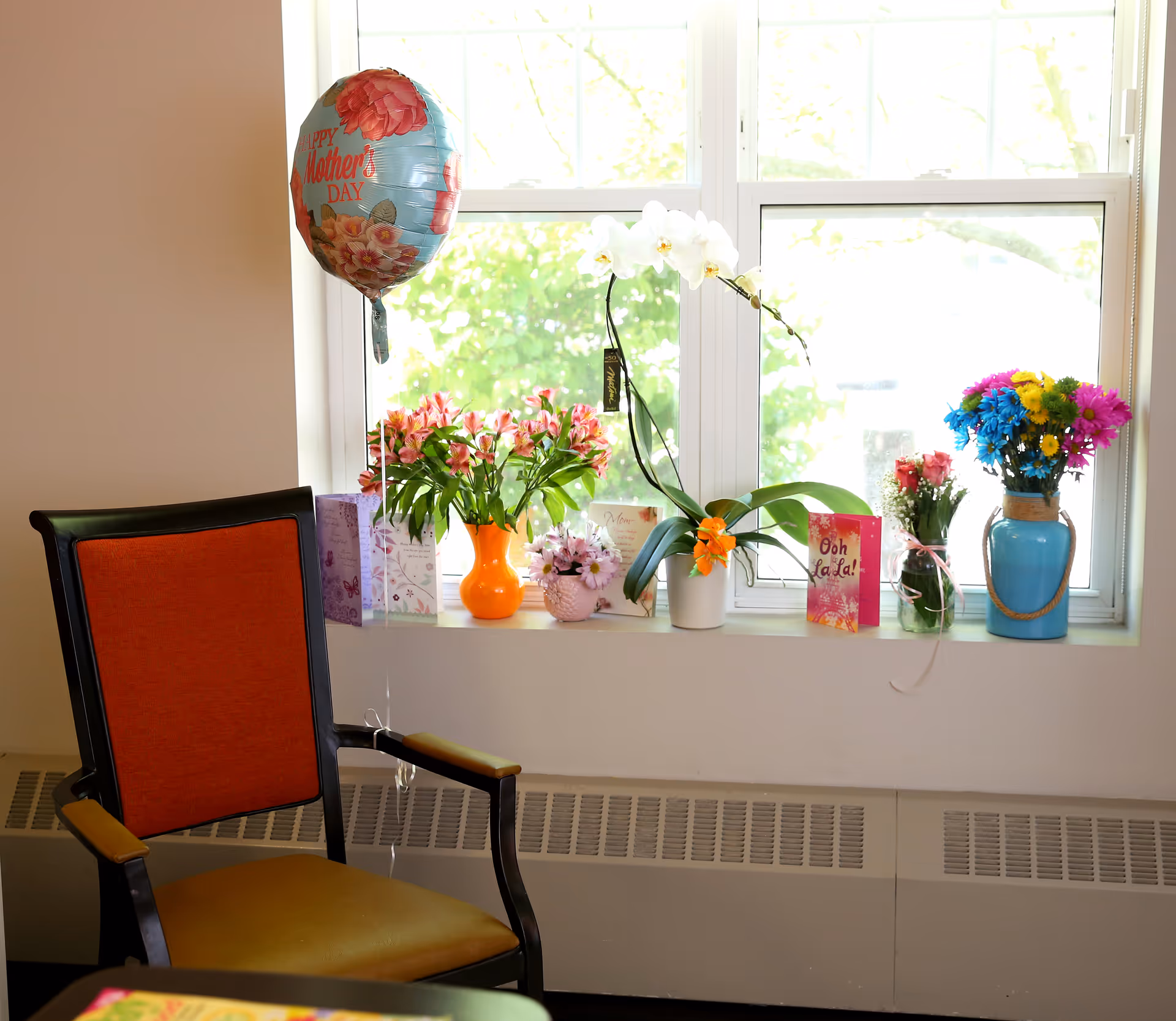 A window sill decorated with various flower arrangements in colorful vases and Mother's Day cards. A balloon with 'Happy Mother's Day' text is tied to a chair with an orange backrest and yellow seat cushion positioned in front of the window.