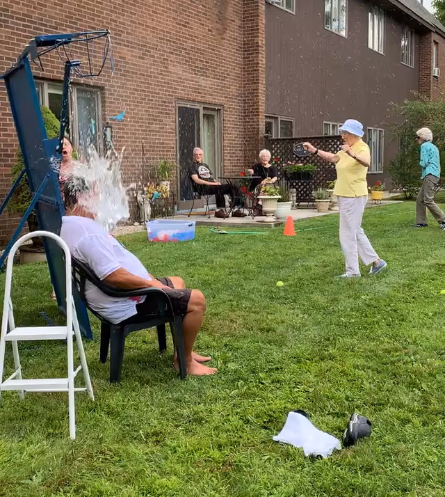 An outdoor scene at a senior living facility where an elderly man is seated on a chair getting splashed with water from a dunk tank. A woman in a yellow shirt and white hat is throwing a ball at the target to dunk him. Other elderly people are sitting and standing nearby, watching the activity on the grass lawn in front of a brick building.