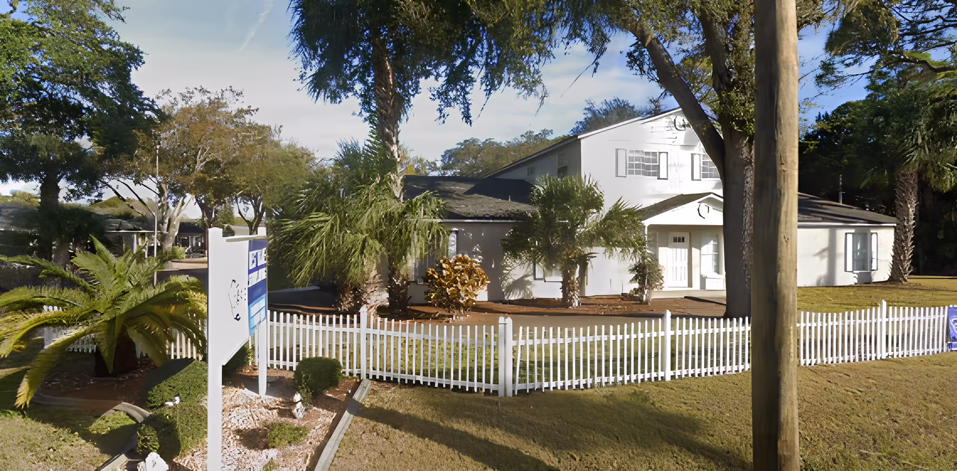 Exterior view of a white senior living facility building surrounded by trees and a white picket fence, with a signpost in the front yard and a clear blue sky above.
