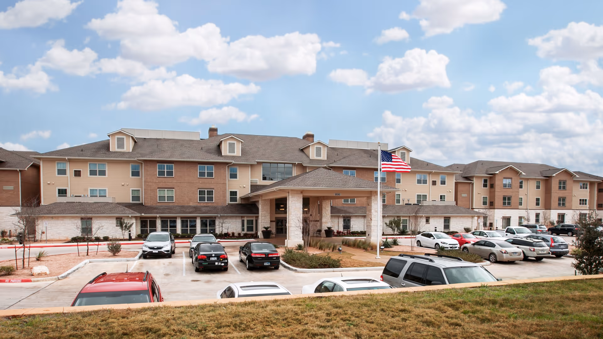 Front exterior view of a large three-story retirement resort building with a covered entrance, multiple windows, an American flag on a flagpole, and a parking lot with several cars in front under a partly cloudy sky.
