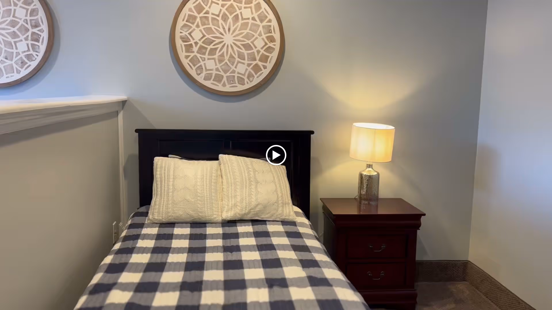 Small bedroom with a single bed dressed in a blue-and-white checkered blanket, two pillows, a dark headboard, a nightstand with a lamp, and round wall decor.
