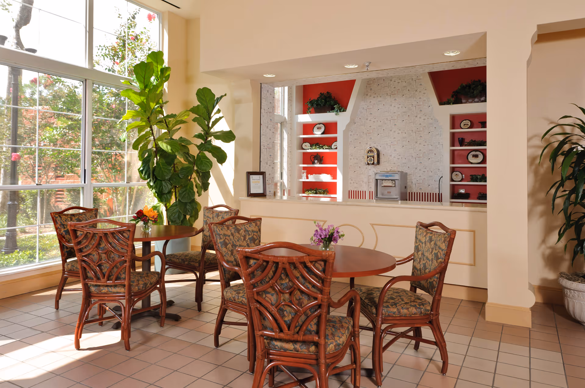 A bright interior room with large windows letting in natural light. There are two round wooden tables surrounded by six cushioned chairs with patterned upholstery. A large green plant is placed near the window, and another plant is in a pot on the right side. The back wall features a recessed area with red shelves displaying decorative plates and plants, and a small appliance is placed on the counter.