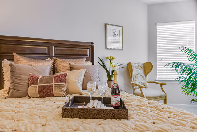 A cozy bedroom with a wooden headboard, multiple decorative pillows on a beige quilted bedspread, a tray with two wine glasses and a bottle on the bed, a potted plant, a framed picture on the wall, a cushioned armchair with floral upholstery, and a window with white blinds letting in natural light.