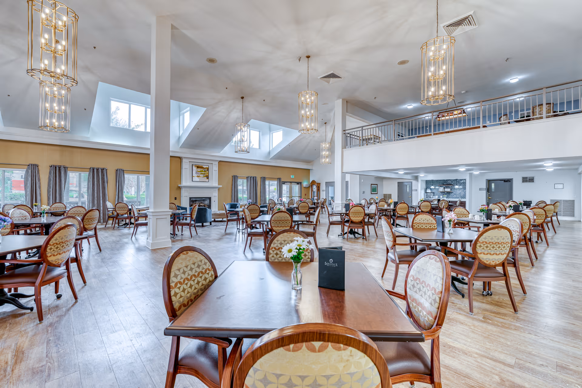 Spacious dining room in a senior living facility with multiple round and square wooden tables surrounded by cushioned chairs. The room features large windows with curtains, a fireplace, high ceilings with skylights, and elegant hanging light fixtures. The floor is wooden, and there is a mezzanine level visible in the background.