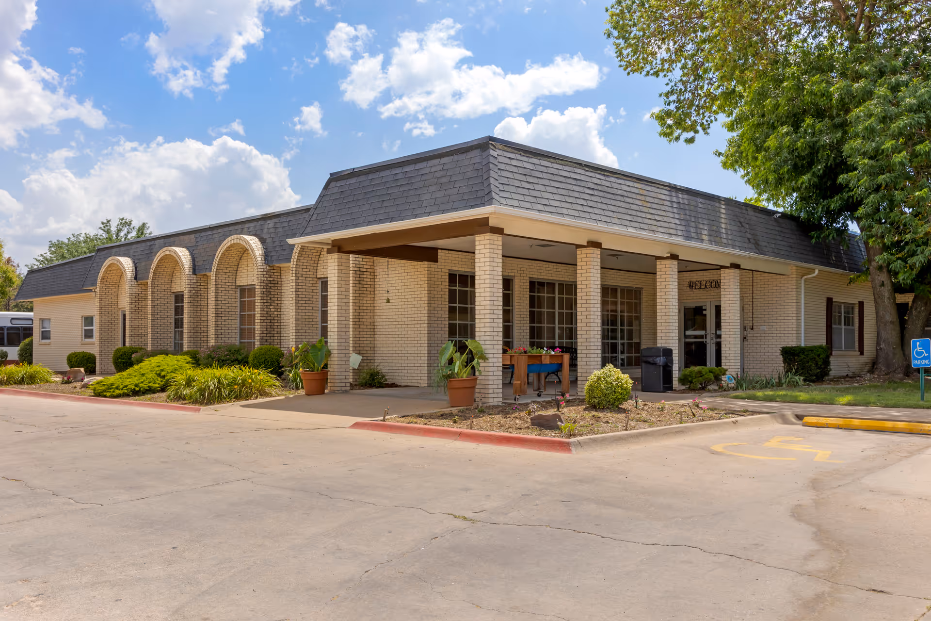 Front exterior of a single-story brick rehabilitation and health care center showing a covered entrance with columns, landscaping, and a parking area.
