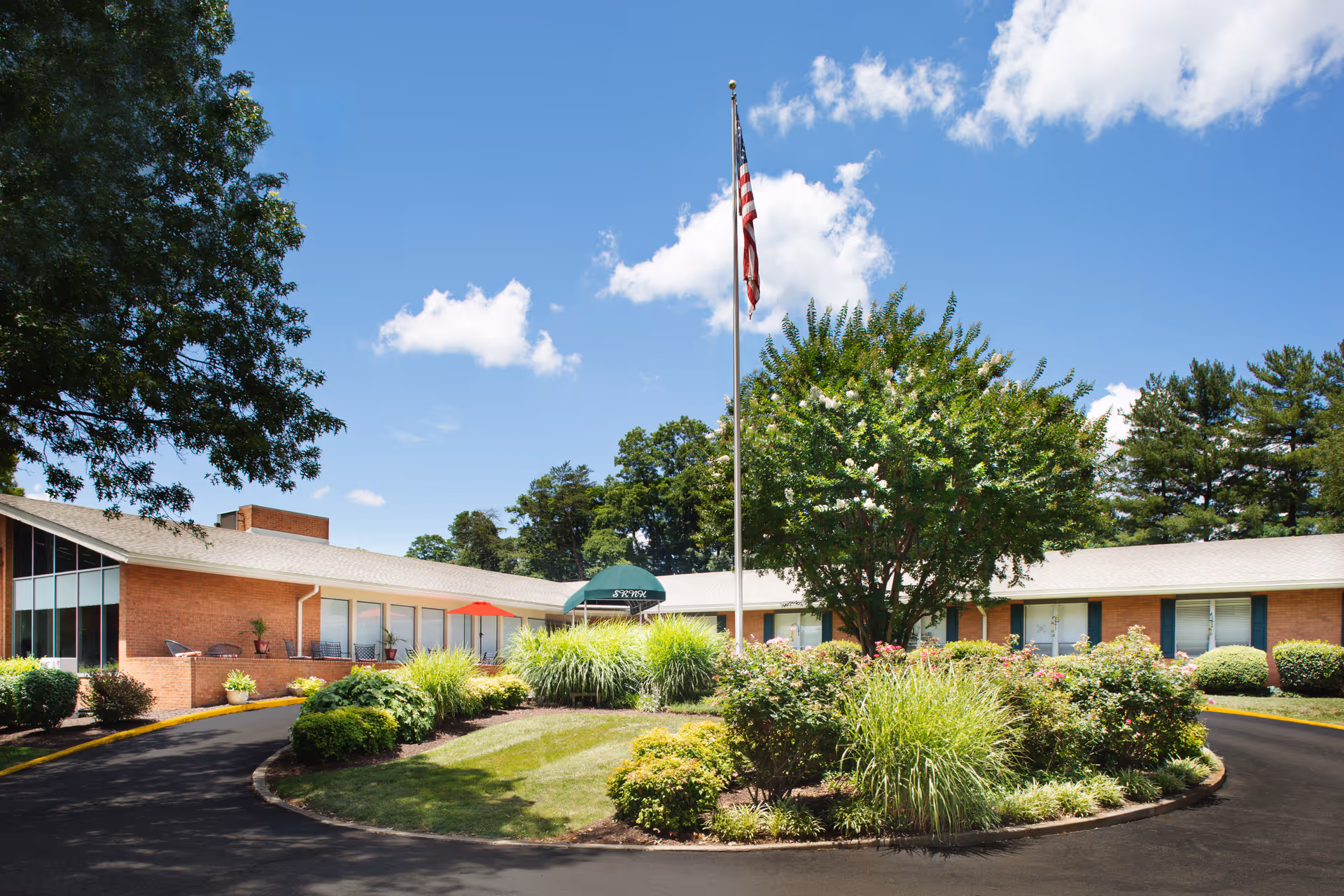 Exterior view of South Roanoke Nursing Home showing a single-story brick building with large windows, surrounded by well-maintained landscaping including bushes and trees. A flagpole with an American flag stands in the center of a circular driveway under a blue sky with scattered clouds.