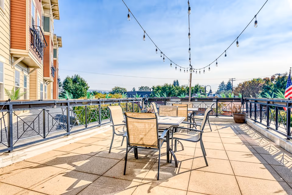 Outdoor patio area at Sellwood Senior Living with several tables and chairs arranged on a tiled floor, string lights hanging above, and a view of trees and buildings in the background under a clear blue sky.