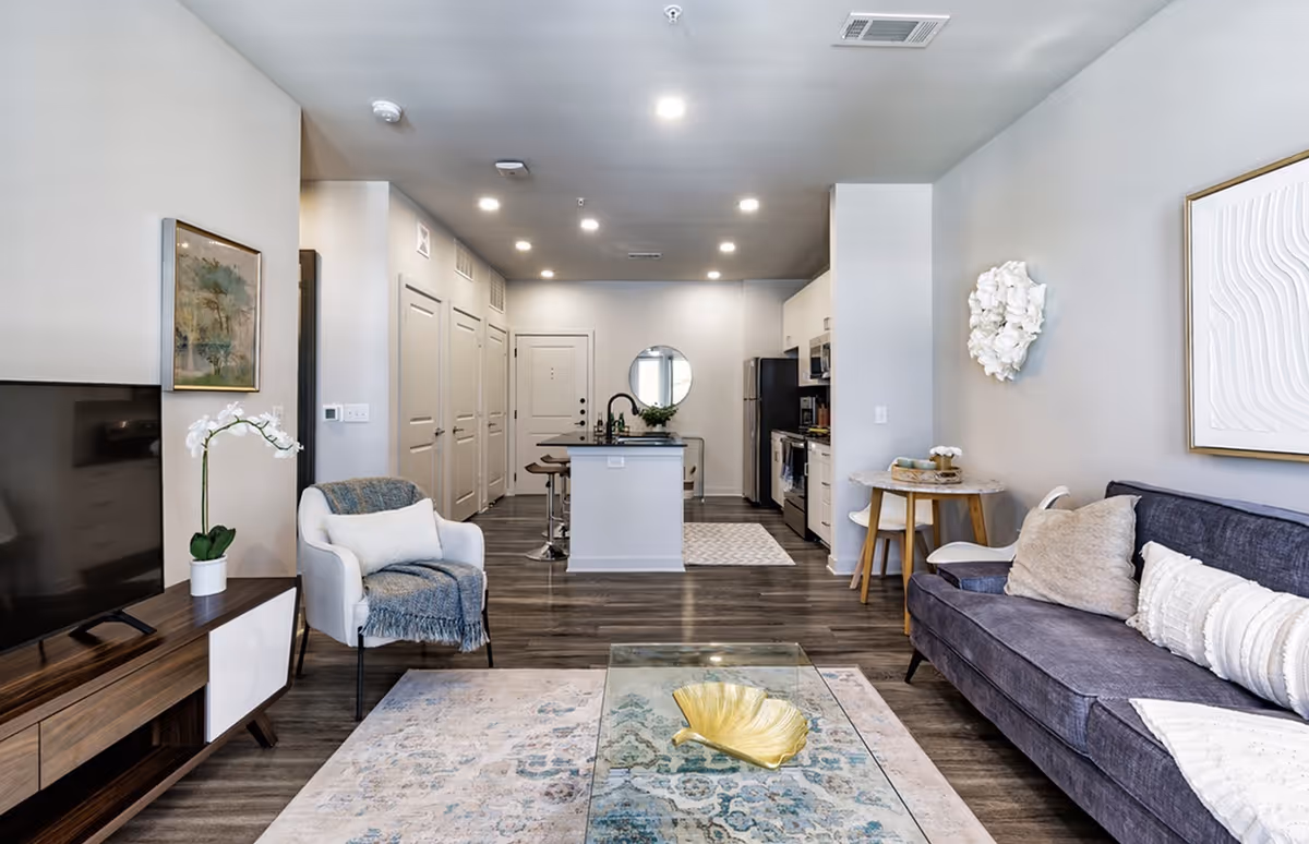 Modern open-concept living area with a dark blue sofa adorned with beige and white pillows on the right, a white armchair with a blue throw blanket on the left, a glass coffee table with a decorative golden leaf centerpiece, and a wooden TV stand with a flat-screen TV and a white orchid plant. The background shows a kitchen with white cabinetry, stainless steel appliances, a kitchen island with bar stools, and a round mirror on the far wall.