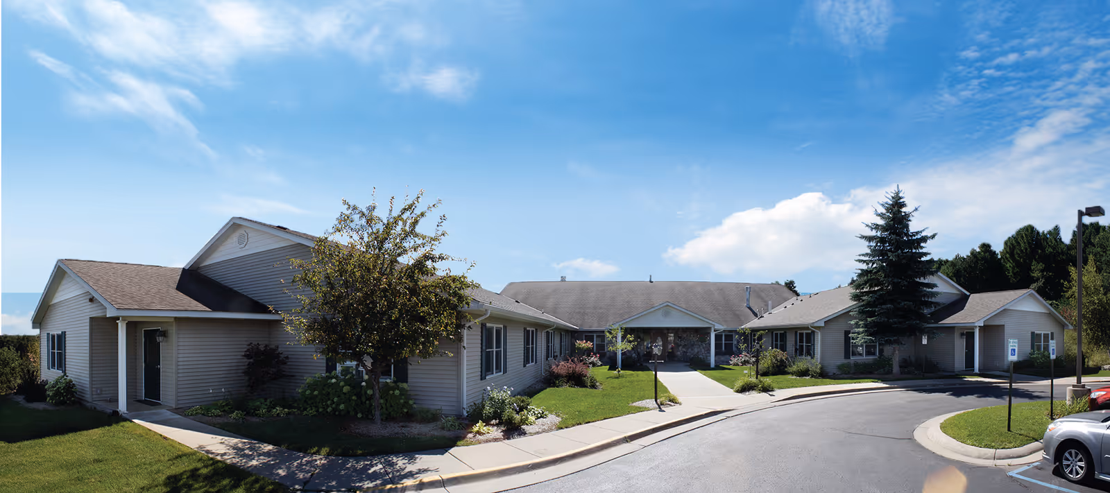 Exterior view of a single-story senior living facility building with beige siding, a gray shingled roof, and landscaped greenery including trees and bushes under a blue sky with some clouds. There is a curved driveway and parking area in front of the building.