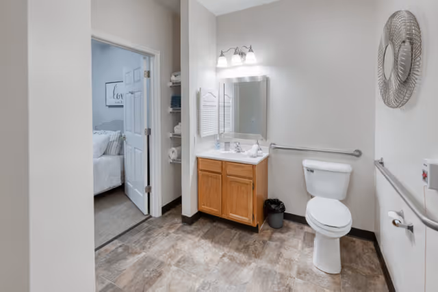 A clean and spacious bathroom with a toilet, a wooden vanity with a sink, a mirror above the sink, and grab bars on the walls. The bathroom has tiled flooring and neutral-colored walls. An open door leads to a bedroom with a bed and wall art visible.