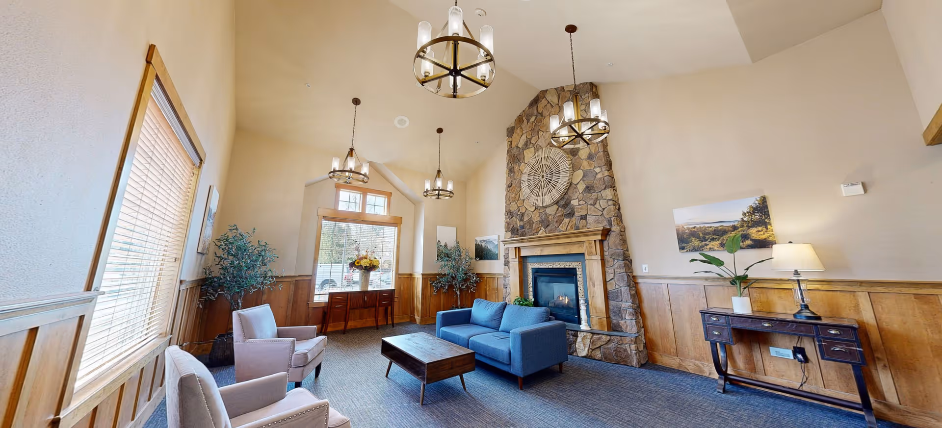 A cozy living room with a high ceiling featuring three hanging light fixtures. The room has a stone fireplace with a decorative piece above it, a blue sofa, two light-colored armchairs, a wooden coffee table, and wooden paneling on the lower half of the walls. There are large windows with blinds, potted plants, and framed landscape pictures on the walls.