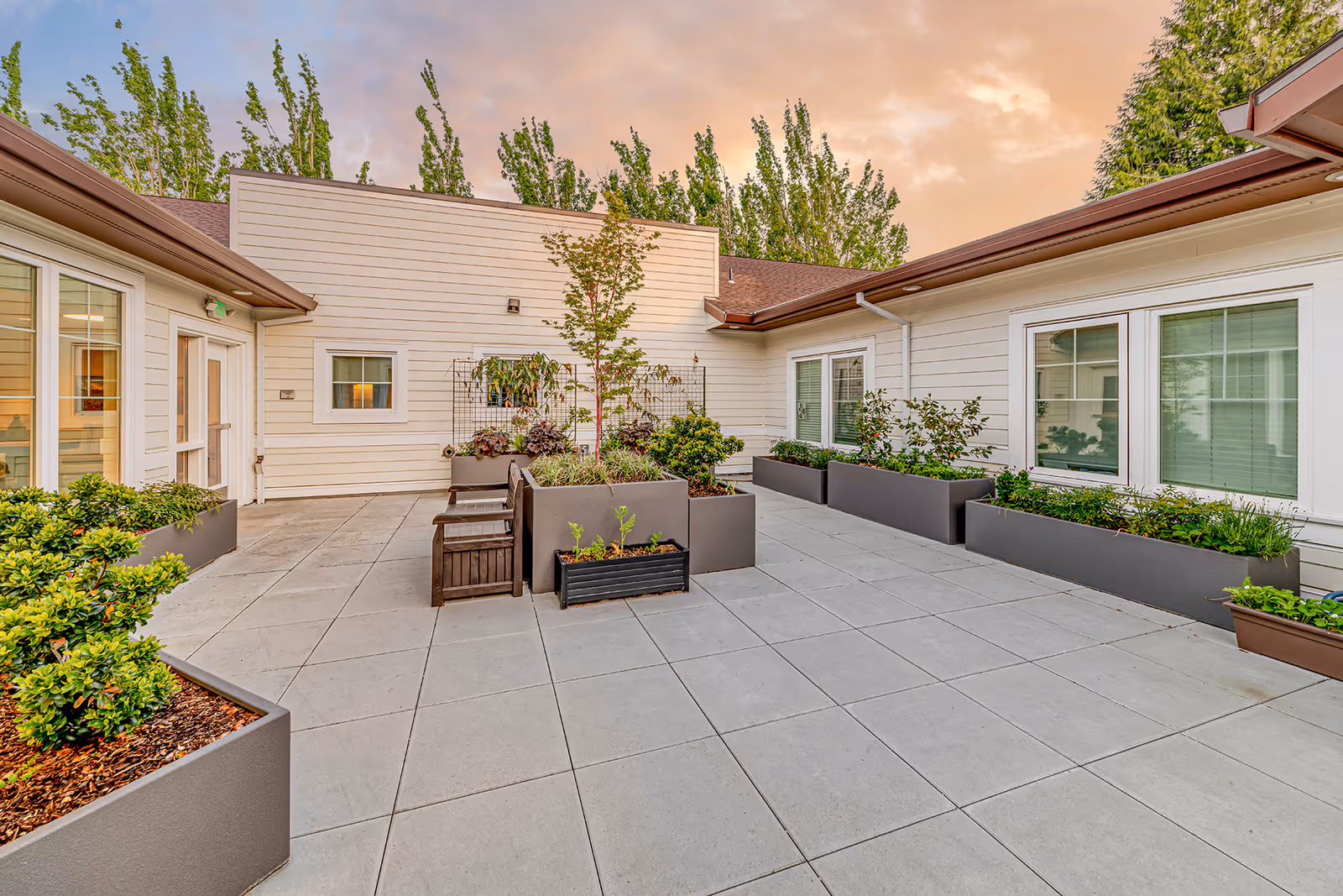 Outdoor courtyard area at Courtyard At Mount Tabor featuring large square tiled flooring, multiple rectangular planters with green plants and small trees, wooden benches, and white building walls with windows surrounding the space under a partly cloudy sky at sunset.