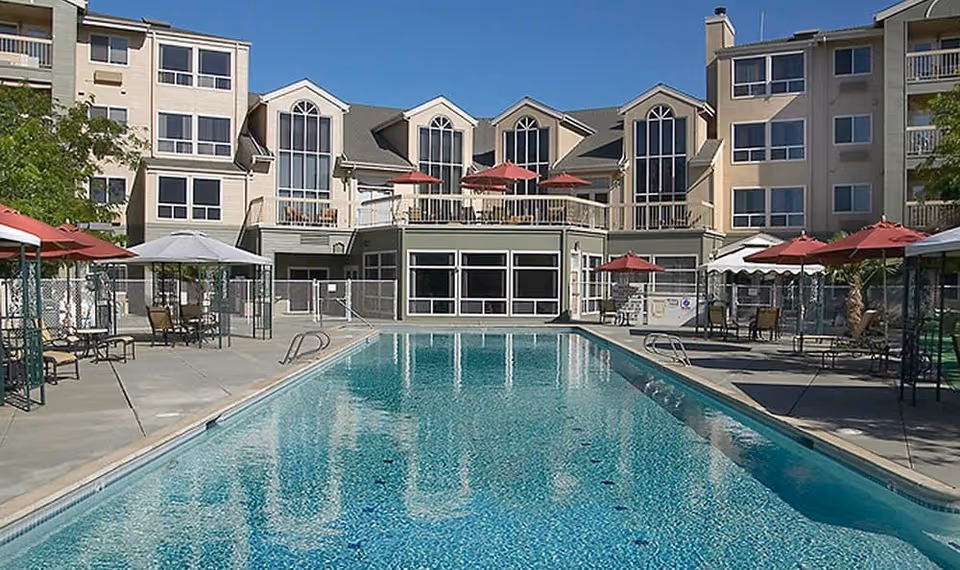 Outdoor swimming pool with clear blue water surrounded by a concrete deck with several tables, chairs, and red and white umbrellas. In the background, there is a multi-story residential building with large windows and balconies.
