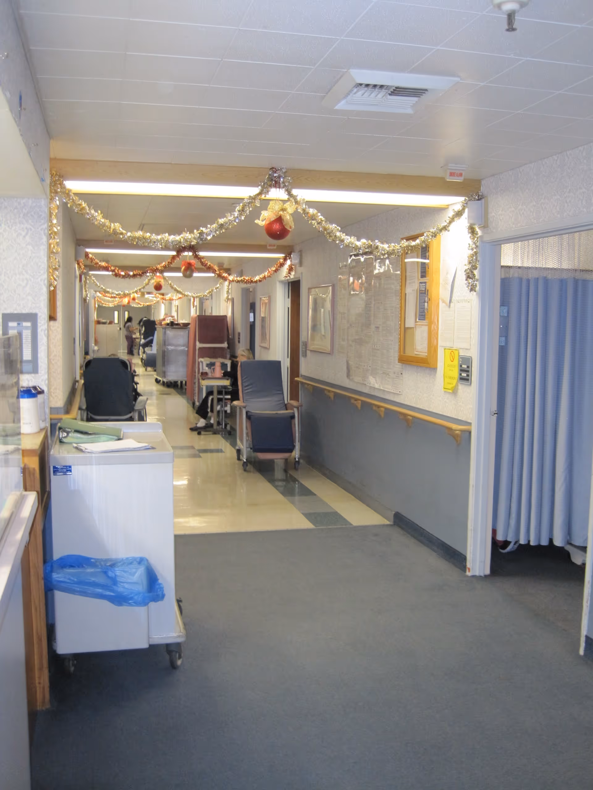A long hallway in a healthcare facility decorated with tinsel and red ornaments hanging from the ceiling. The hallway has chairs and medical equipment along the walls, with a blue curtain partially visible on the right side. The floor is a combination of carpet and linoleum tiles, and there are handrails on the walls.
