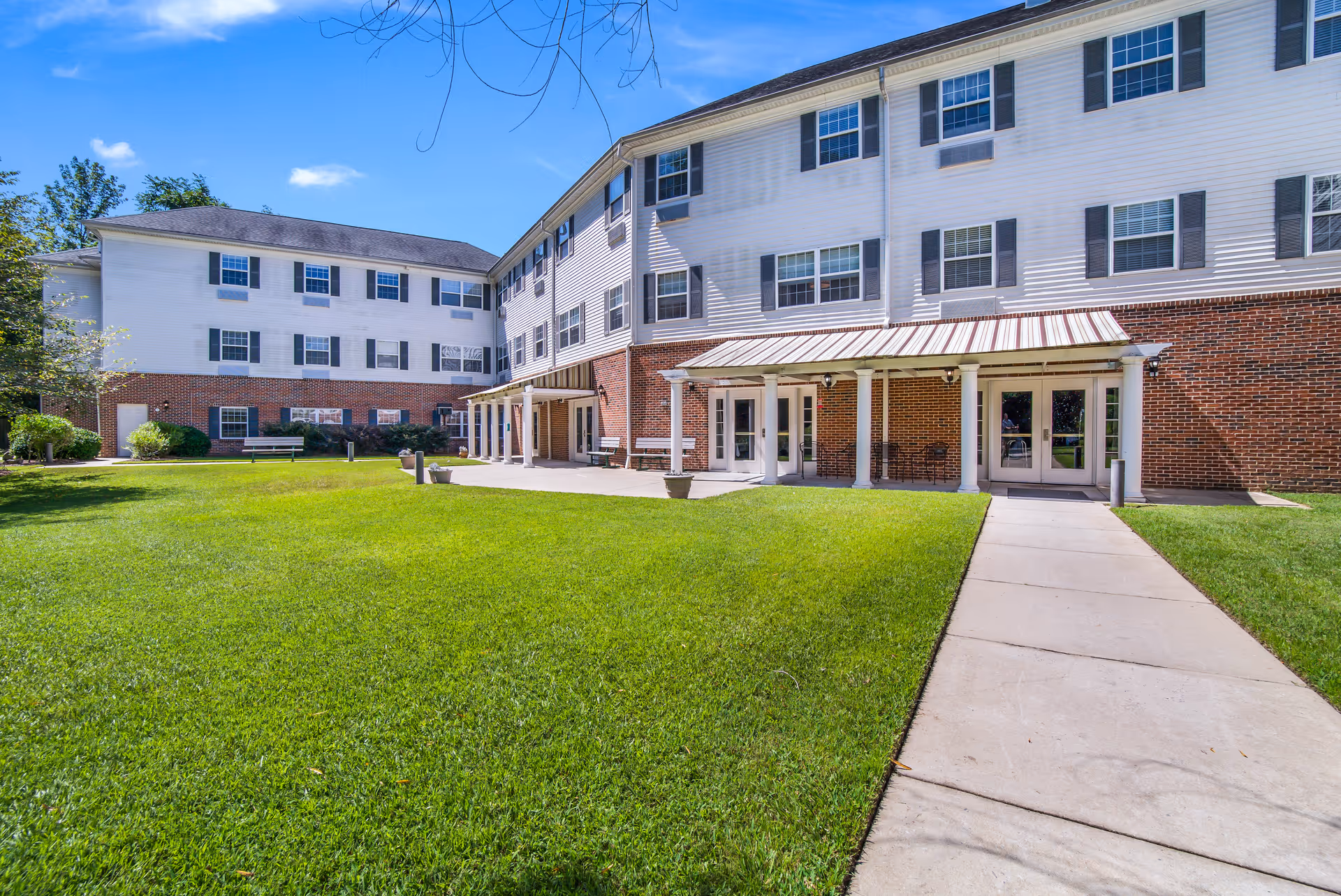 Exterior view of a multi-story senior living facility building with white siding and red brick lower walls. The building has multiple windows with black shutters and a covered entrance supported by white columns. There is a well-maintained green lawn and a concrete walkway leading to the entrance under a clear blue sky.